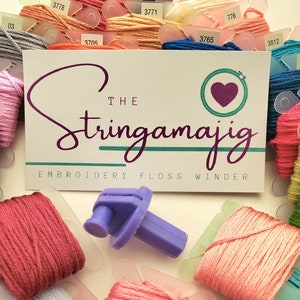 Può includere: Una collezione di fili da ricamo colorati e un avvolgitore di filo da ricamo viola. Al centro c'è una scheda con il testo "The Stringamajig Embroidery Floss Winder". Il filo è in varie tonalità di rosa, arancione e blu.