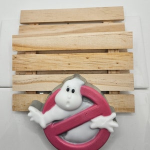 May include: A pink and white soap bar shaped like the Ghostbusters logo with a ghost inside. The soap is on a wooden soap dish.