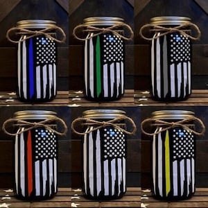 May include: Six mason jars decorated with an American flag design. Each jar has a different colored stripe down the center, representing different first responder groups: blue for police, green for military, gray for firefighters, red for EMT, yellow for dispatch, and white for all first responders.
