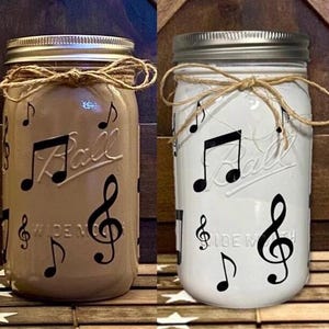 May include: Two mason jars decorated with black musical notes. One jar is brown and the other is white. Both jars have a brown twine bow around the lid.