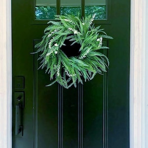 May include: A circular wreath composed of lush green foliage and small white flowers hanging on a dark forest green front door. The door features a rectangular glass window pane at the top and a black metal handle set on the left side.