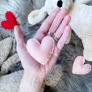 May include: A hand holding a pink felt heart with white stitching. There are two other felt hearts in the background, one red and one pink.