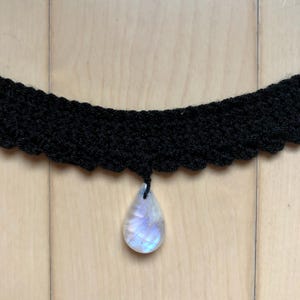 May include: A black crocheted choker necklace with a teardrop-shaped iridescent moonstone pendant.