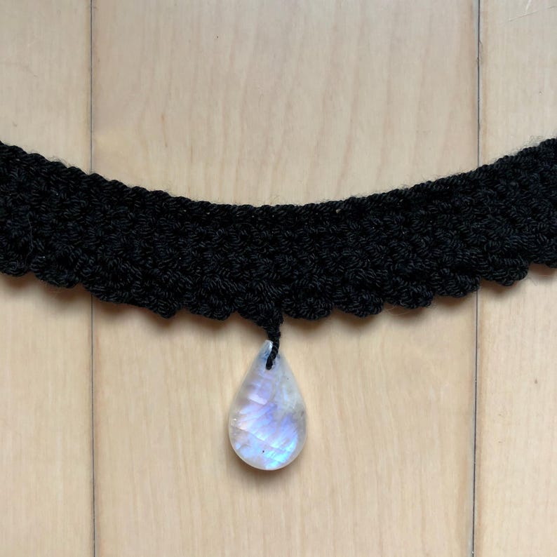 May include: A black crocheted choker necklace with a teardrop-shaped iridescent moonstone pendant.