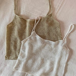 Op de afbeelding: Twee gebreide tanktops, één in lichtbeige en de andere in wit. Beide tops hebben dunne spaghettibandjes en een ronde halslijn. De tops zijn gemaakt van een getextureerde gebreide stof en liggen op een wit oppervlak.