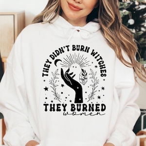 May include: White hoodie with a black graphic design. The design features a hand holding a crystal, a crescent moon, and the text "THEY DIDN'T BURN WITCHES THEY BURNED WOMEN". The hoodie has a drawstring hood and a kangaroo pocket.