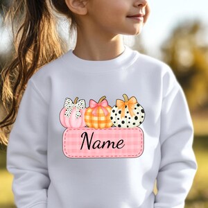 May include: White sweatshirt featuring a graphic of three pumpkins with bows and a pink name tag. The pumpkins have different patterns: polka dots, plaid, and a solid color. The word "Name" is written on the tag.