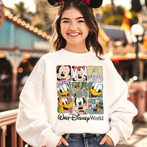 Op de afbeelding: Witte trui met een kleurrijke afbeelding van verschillende Disney-figuren, waaronder Mickey Mouse, Donald Duck en Goofy. De afbeelding is in een rasterpatroon gerangschikt en bevat de tekst "Walt Disney World".