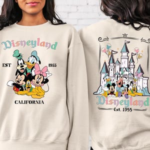 May include: A beige sweatshirt with a colourful Disneyland graphic on the front and back. The front graphic features Mickey Mouse, Minnie Mouse, Goofy, Pluto, and Donald Duck. The back graphic features a colourful illustration of Sleeping Beauty's castle with Mickey Mouse, Minnie Mouse, Goofy, Pluto, and Donald Duck in front of it. The text "Disneyland Est. 1955" is printed on both the front and back of the sweatshirt.