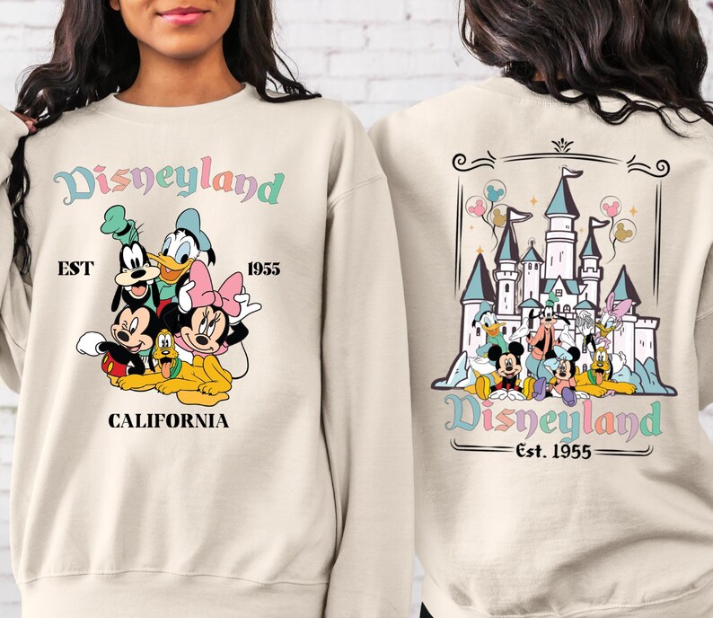 May include: A beige sweatshirt with a colourful Disneyland graphic on the front and back. The front graphic features Mickey Mouse, Minnie Mouse, Goofy, Pluto, and Donald Duck. The back graphic features a colourful illustration of Sleeping Beauty's castle with Mickey Mouse, Minnie Mouse, Goofy, Pluto, and Donald Duck in front of it. The text "Disneyland Est. 1955" is printed on both the front and back of the sweatshirt.