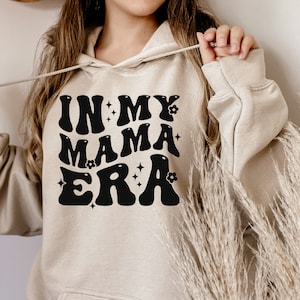 Pu&ograve; includere: Una felpa con cappuccio beige con una grafica nera che dice "In My Mama Era" in un font retr&ograve; con fiori e stelle.