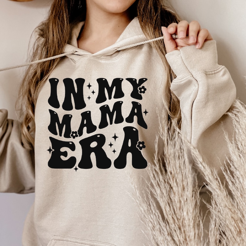Pu&ograve; includere: Una felpa con cappuccio beige con una grafica nera che dice "In My Mama Era" in un font retr&ograve; con fiori e stelle.