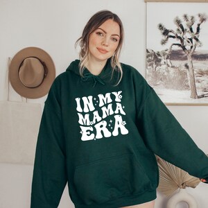Pu&ograve; includere: Una felpa con cappuccio verde scuro con il testo "In My Mama Era" in bianco con un font retr&ograve; e stelle bianche.