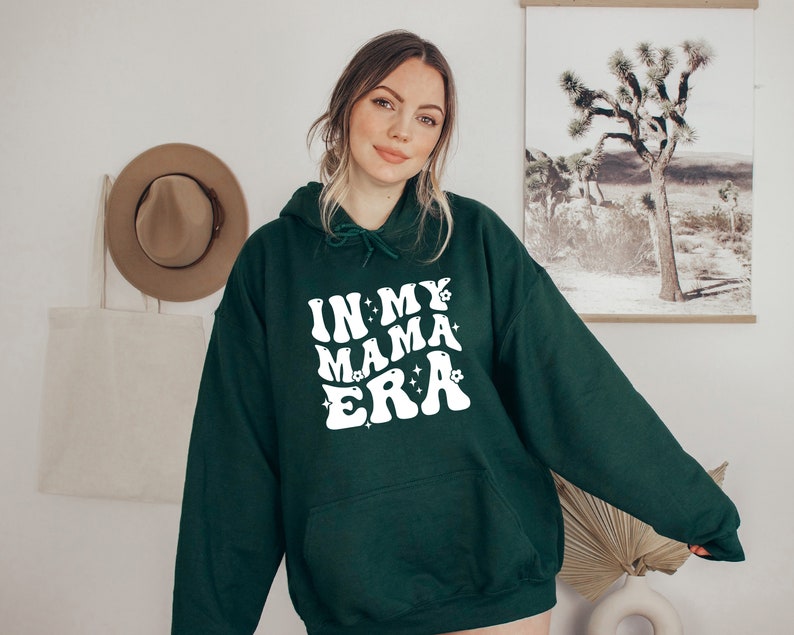 Pu&ograve; includere: Una felpa con cappuccio verde scuro con il testo "In My Mama Era" in bianco con un font retr&ograve; e stelle bianche.