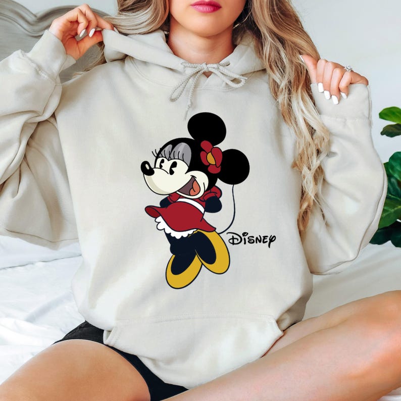 Puede incluir: Una sudadera con capucha gris claro con una imagen de dibujos animados de Minnie Mouse con un vestido rojo y zapatos amarillos. El texto "DISNEY" est&aacute; impreso debajo de la imagen.