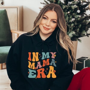 Pu&ograve; includere: Una felpa con cappuccio nera con una grafica colorata che dice "In My Mama Era" con stelle e fiori. La felpa &egrave; indossata da una persona con i capelli lunghi e biondi.