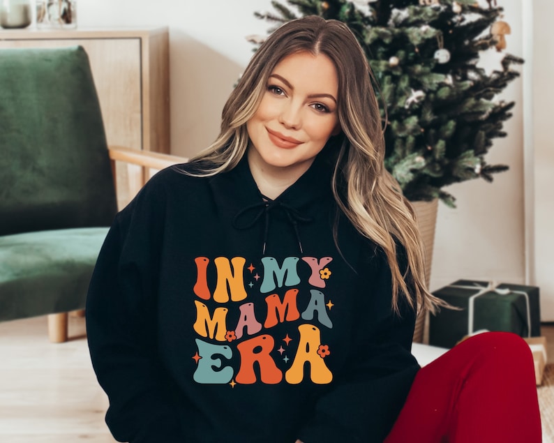 Pu&ograve; includere: Una felpa con cappuccio nera con una grafica colorata che dice "In My Mama Era" con stelle e fiori. La felpa &egrave; indossata da una persona con i capelli lunghi e biondi.
