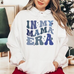Pu&ograve; includere: Felpa con cappuccio bianca con il testo "In My Mama Era" in lettere retr&ograve; blu e viola con fiori e stelle.