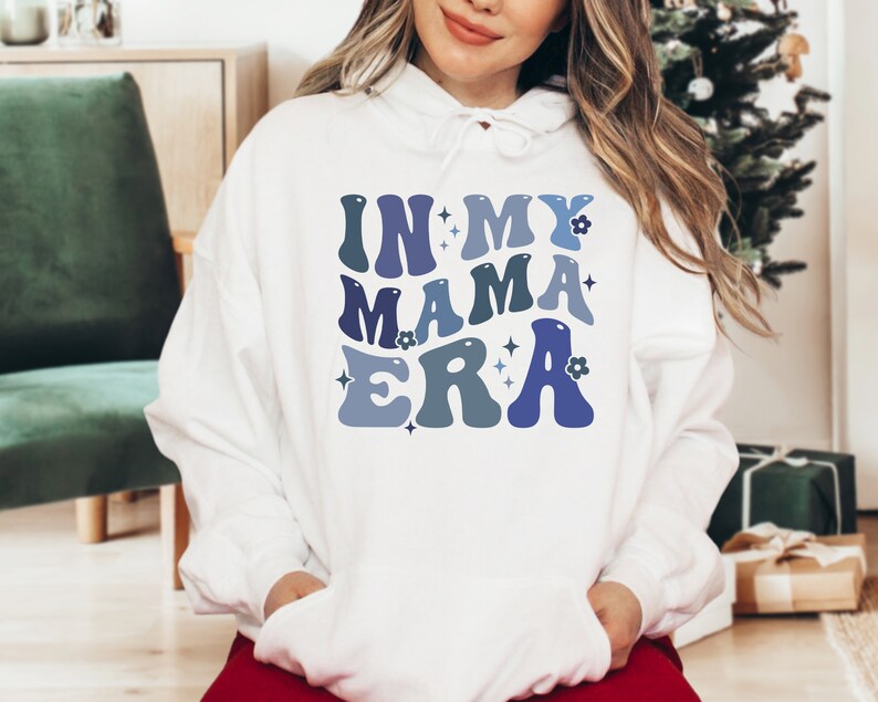 Pu&ograve; includere: Felpa con cappuccio bianca con il testo "In My Mama Era" in lettere retr&ograve; blu e viola con fiori e stelle.