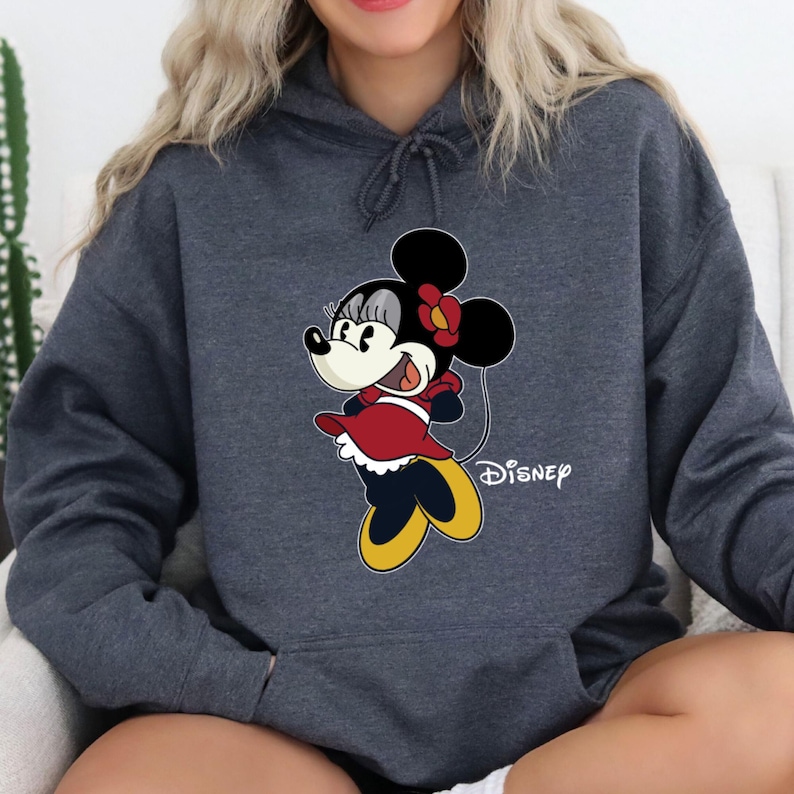 Puede incluir: Una sudadera con capucha gris oscuro con una imagen de dibujos animados de Minnie Mouse con un vestido rojo y zapatos amarillos. El texto "DISNEY" est&aacute; impreso en la esquina inferior derecha de la sudadera.