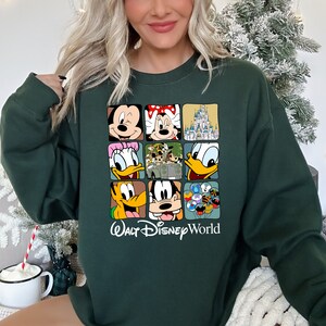 Op de afbeelding: Een donkergroen sweatshirt met een grafische print van verschillende Disney-figuren, waaronder Mickey Mouse, Minnie Mouse, Donald Duck, Goofy en Pluto. De grafiek bevat ook het iconische kasteel van Assepoester en de tekst "Walt Disney World".