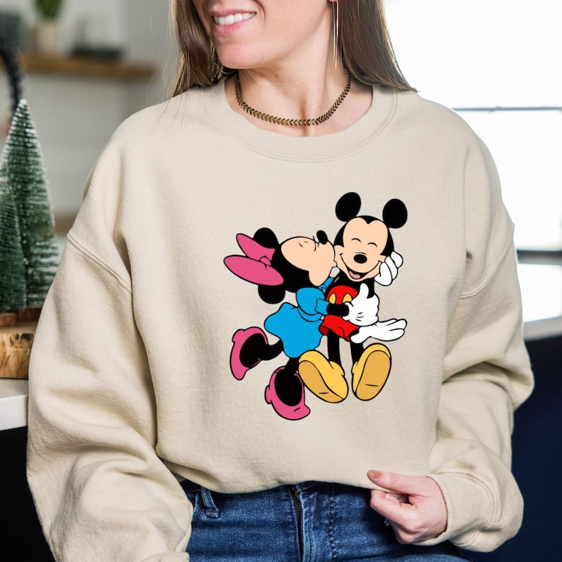 May include: Beige sweatshirt featuring a graphic of Mickey Mouse and Minnie Mouse embracing. Mickey is wearing red shorts, a black shirt, and yellow shoes. Minnie is wearing a blue dress, pink shoes, and a pink bow.