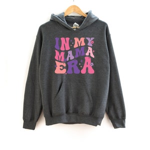 Pu&ograve; includere: Una felpa con cappuccio grigio scuro con il testo "IN MY MAMA ERA" stampato in lettere rosa, viola e rosa chiaro con un font retr&ograve;. Le lettere sono contornate di bianco.