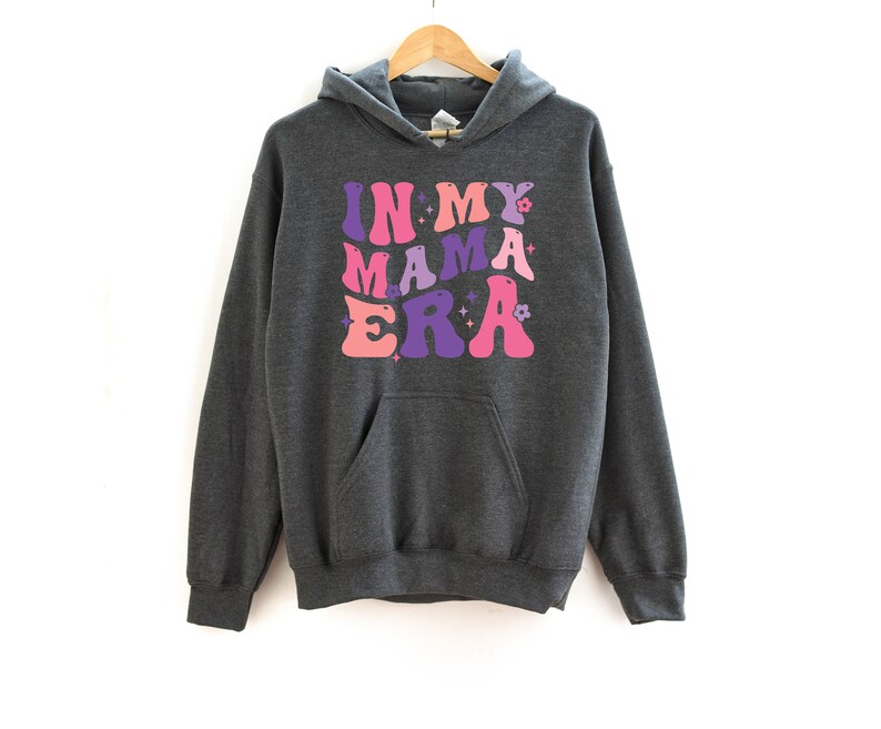 Pu&ograve; includere: Una felpa con cappuccio grigio scuro con il testo "IN MY MAMA ERA" stampato in lettere rosa, viola e rosa chiaro con un font retr&ograve;. Le lettere sono contornate di bianco.