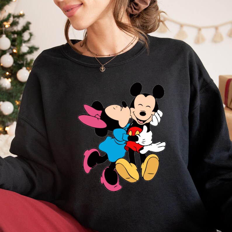May include: Black sweatshirt featuring a colorful graphic of Mickey Mouse and Minnie Mouse embracing. Minnie Mouse is wearing a blue dress and pink bow, while Mickey Mouse wears red shorts. The design is a classic cartoon style.