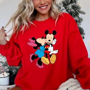 May include: A red sweatshirt featuring a graphic of Mickey Mouse and Minnie Mouse embracing. Mickey Mouse is wearing red shorts, and Minnie Mouse is wearing a blue dress and pink bow. The sweatshirt is a solid red color.
