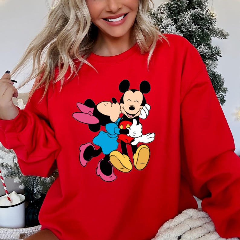 May include: A red sweatshirt featuring a graphic of Mickey Mouse and Minnie Mouse embracing. Mickey Mouse is wearing red shorts, and Minnie Mouse is wearing a blue dress and pink bow. The sweatshirt is a solid red color.