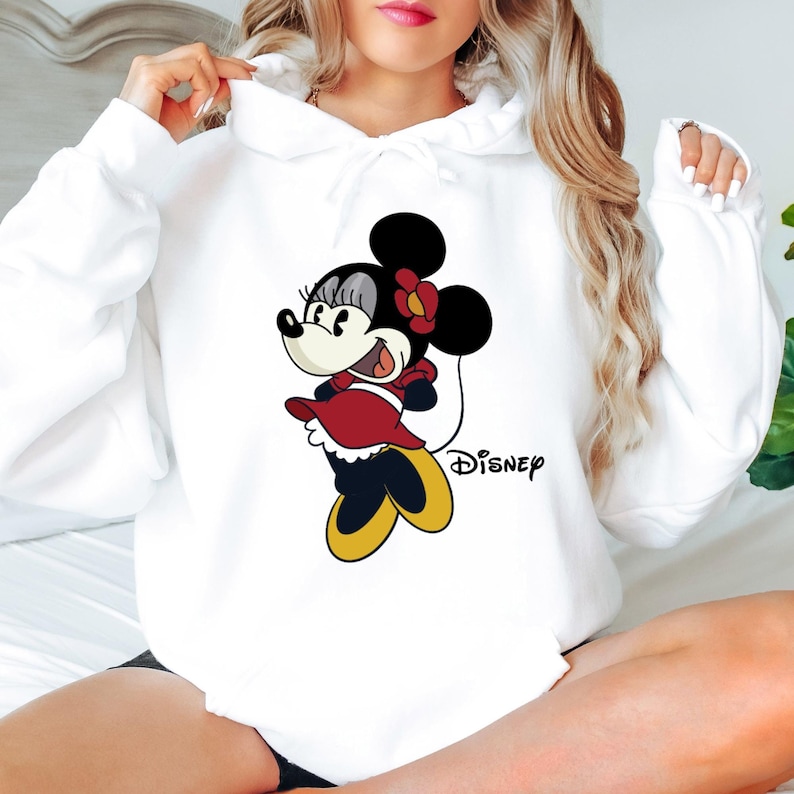 Puede incluir: Sudadera con capucha blanca con una imagen de dibujos animados de Minnie Mouse con un vestido rojo y zapatos amarillos. El texto "DISNEY" est&aacute; impreso debajo de la imagen.