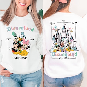 May include: White sweatshirt with a colourful Disneyland graphic featuring Mickey Mouse, Minnie Mouse, Donald Duck, Goofy, and Pluto. The front of the sweatshirt says "Disneyland EST 1955 CALIFORNIA" and the back of the sweatshirt says "Disneyland Est. 1955" with a colourful illustration of Sleeping Beauty's castle.