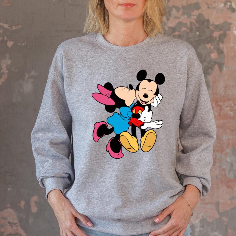 May include: A gray sweatshirt featuring a colorful graphic of Mickey Mouse and Minnie Mouse embracing. Minnie is wearing a blue dress and pink bow and shoes. Mickey is wearing red shorts and yellow shoes.