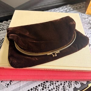May include: A dark brown velvet clutch bag featuring a curved gold-tone metal frame with a kiss-lock closure. The soft fabric has a slight sheen and a folded design, showcasing a smooth texture across the exterior surface of the accessory.