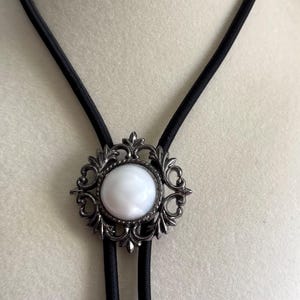 May include: A bolo tie featuring a white, oval-shaped stone set in an ornate, silver-toned metal frame. The frame has a detailed, swirling design. The tie has a black cord.