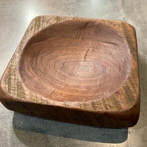 May include: A square wooden bowl with a rounded interior. The bowl is made of dark brown wood with a natural grain pattern.