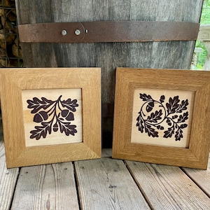 May include: Two square wooden frames with light-colored wood centers featuring dark brown laser-cut designs of oak leaves and acorns. Each frame has a natural wood grain finish and a beveled edge, displaying intricate botanical patterns.