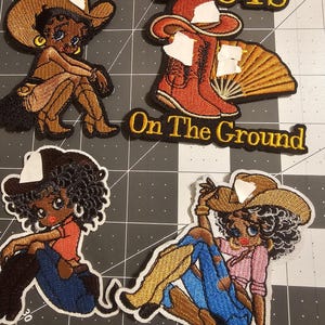 Puede incluir: Parches bordados con un tema de vaquera de dibujos animados. Un parche tiene el texto "BOOTS On The Ground". Los parches presentan una vaquera con sombrero y botas de vaquero, en tonos marrones, beige, rojos, azules y rosas.