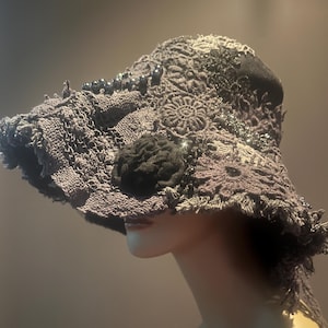 May include: A dark gray, wide-brimmed hat with a textured, woven design. The hat features a black fabric flower and is embellished with dark beads and sparkling accents. The brim has a frayed edge, adding a unique, handcrafted look.