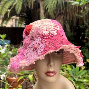 May include: A pink crochet hat with a wide brim. The hat features various shades of pink, with a textured design and a red flower accent. The brim has a frayed edge. The hat is likely handmade.