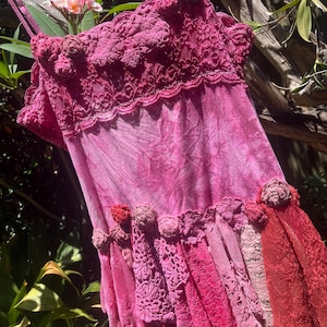 Puede incluir: Vestido magenta con efecto tie-dye. La parte superior presenta detalles de encaje intrincados y adornos florales. La parte inferior del vestido tiene capas de encaje y tela en varios tonos de rosa y rojo. El vestido está colgado al aire libre.