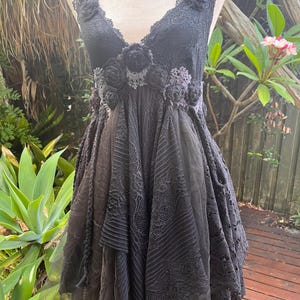 May include: Black sleeveless dress with a V-neckline, adorned with lace and floral embellishments. The dress features a layered skirt with varying textures and patterns, creating a unique and elegant design.