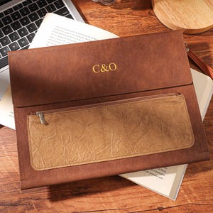 May include: A brown leather clutch bag with a front pocket and a wrist strap. The initials "C&O" are in gold. The bag rests on a book and near a laptop on a wooden surface. The clutch is approximately 30 cm long.