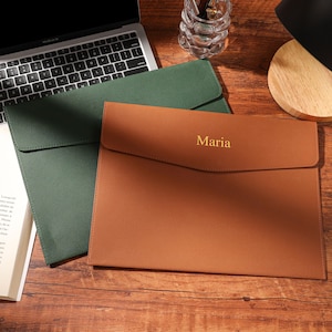 Puede incluir: Dos fundas de documentos de cuero en verde y marrón, con un portátil y un libro en el fondo. La funda marrón tiene el nombre "Maria" grabado en dorado. Los artículos están sobre una superficie de madera.