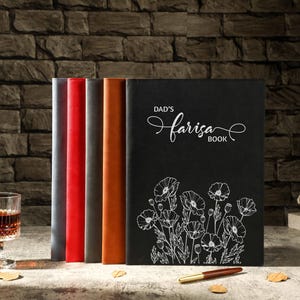 Könnte beinhalten: Eine Sammlung ledergebundener Notizbücher in verschiedenen Farben, darunter Rot, Grau und Braun. Ein schwarzes Notizbuch zeigt den Text "DAD'S Farisa BOOK" und ein Blumendesign. Ein Stift und ein Glas bernsteinfarbener Flüssigkeit sind ebenfalls vorhanden.