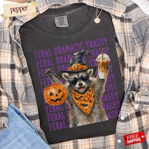 Puede incluir: Una camiseta gris carbón con el gráfico de un mapache con sombrero de bruja, gafas de sol en forma de corazón y un pañuelo naranja con murciélagos. El mapache sostiene un cubo de calabaza y un café helado. El texto FERAL DRAMATIC TRASHY se repite.