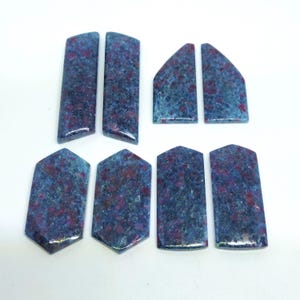 May include: Eight cabochon gemstones with a blue and red speckled pattern. The stones are various shapes and sizes, including rectangular, hexagonal, and triangular.