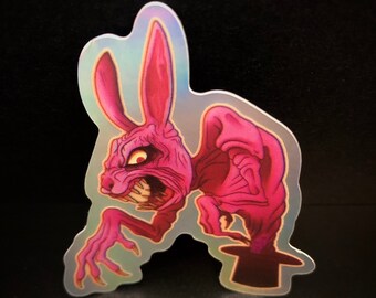May include: A pink rabbit with sharp teeth and claws emerges from a black top hat. The rabbit has a menacing expression and is depicted in a cartoon style. The sticker is holographic and has a glossy finish.