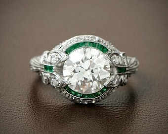 May include: A silver engagement ring with a large round diamond surrounded by a halo of smaller diamonds and a band of emerald green stones.